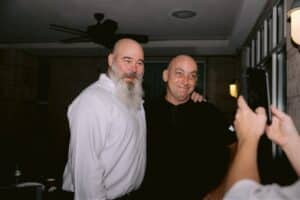 Two men posing for a selfie indoors; the man on the left wears a white shirt and has a long gray beard, the man on the right wears a black shirt.