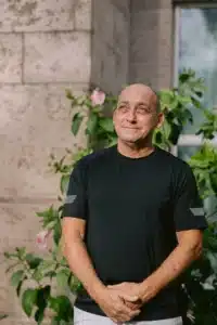 Smiling, bald man in a black T-shirt stands outdoors near a stone wall and green plants.